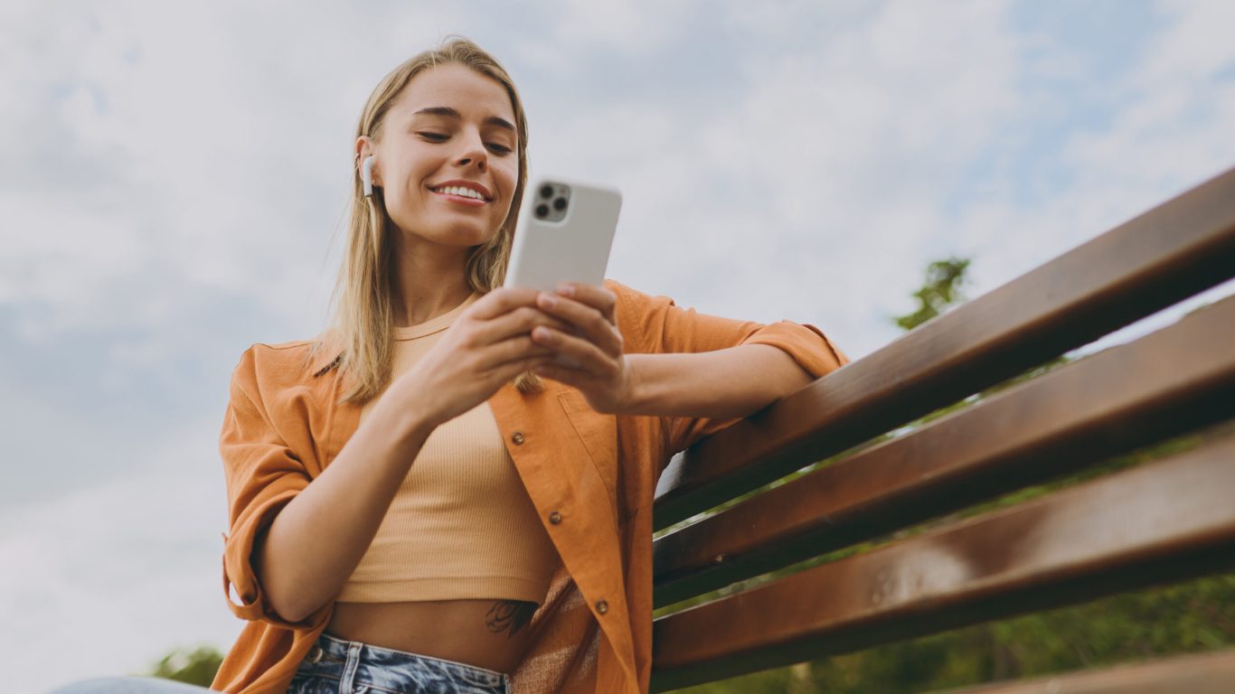 Bottom view young woman wear orange shirt casual clothes use mobile cell phone listen music in earphones sit on bench walk rest relax in spring city park outdoors on nature. Urban lifestyle concept
